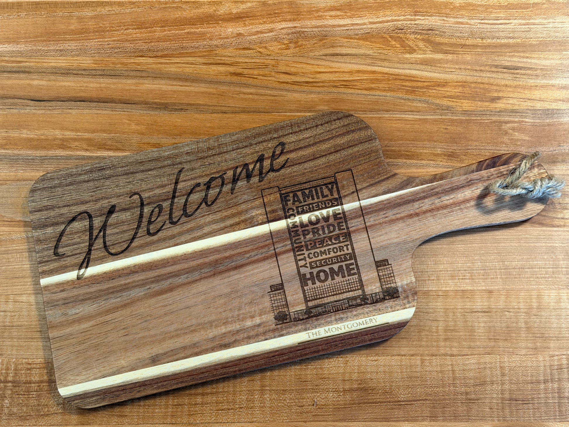 Acacia wood cutting board engraved with the words "Welcome" or "Welcome Home" accompanied by an outline sketch of the recipient's home or apartment/condo building.