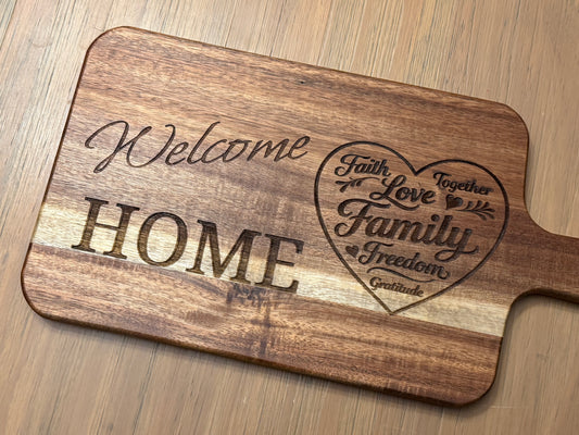 Acacia wood cutting board engraved with the words "Welcome Home" accompanied by a heart design which contains words like "Family", "Faith" and "Love".