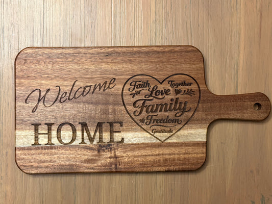 Acacia wood cutting board engraved with the words "Welcome Home" accompanied by a heart design which contains words like "Family", "Faith" and "Love".